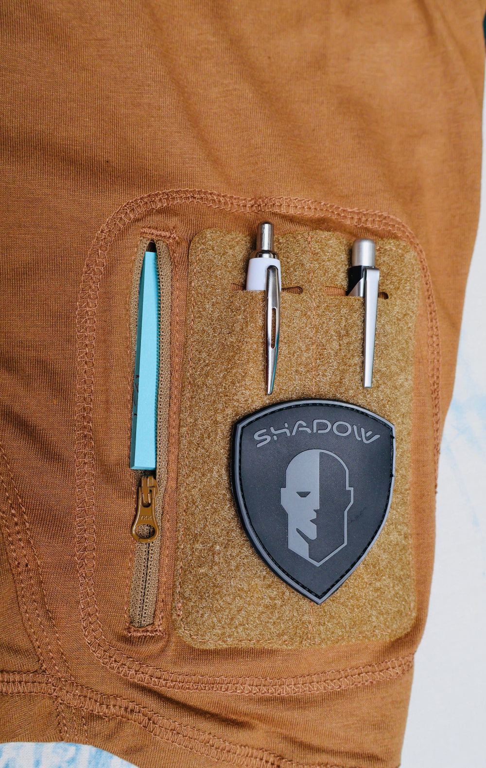 Close-up of a brown tactical vest with a 'Shadow' brand patch and tools.